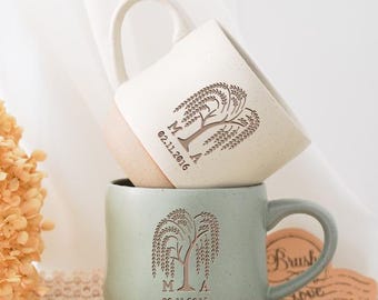 May include: Two ceramic mugs stacked on top of each other. The top mug is off-white with a light brown base, and the bottom mug is a muted green with a light brown base. Both mugs feature a tree design and the date 02.11.2016.