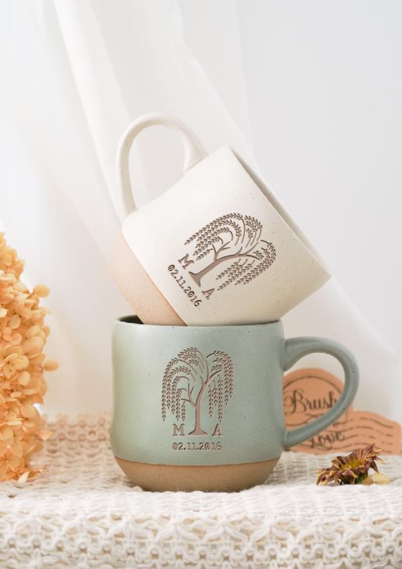 May include: Two ceramic mugs, one stacked on the other. The top mug is off-white with a light brown base, and the bottom mug is a muted green with a light brown base. Both mugs feature a tree design and the date 02.11.2016.