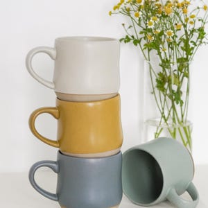 May include: Three ceramic mugs stacked on top of each other. The top mug is white, the middle mug is yellow, and the bottom mug is blue. A fourth mug is light blue and is sitting on a white surface next to a white keyboard.