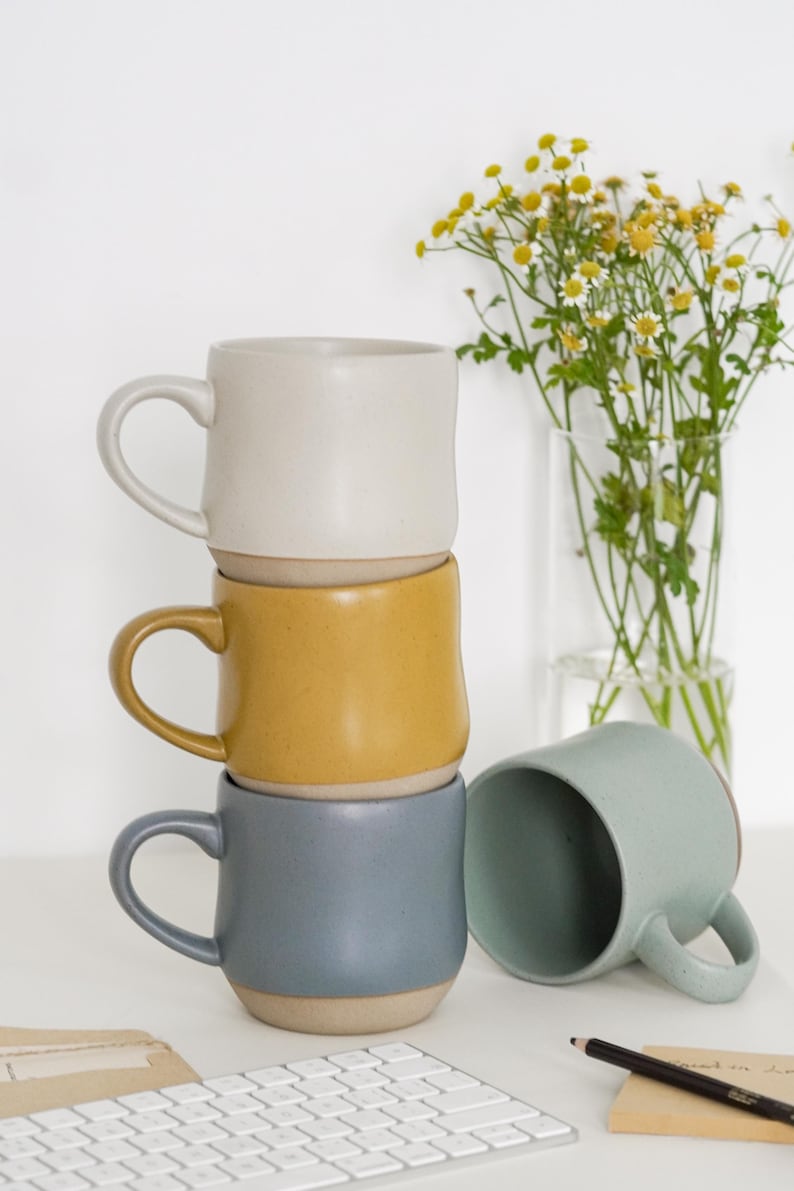 May include: Three ceramic mugs stacked on top of each other. The top mug is white, the middle mug is yellow, and the bottom mug is blue. A fourth mug is light blue and is sitting on a white surface next to a white keyboard.