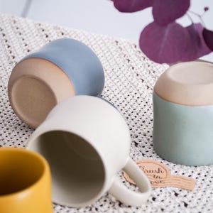 May include: Several ceramic mugs in shades of blue, white, yellow, and mint green are displayed. They are placed on a white crocheted surface, with a purple leaf and a tag that reads "Brush in Love".