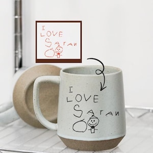 Puede incluir: Taza de cerámica con cuerpo blanco y base marrón. La taza presenta la frase manuscrita "I LOVE SATAN" con una ilustración sencilla. Un platillo a juego es visible en el fondo.