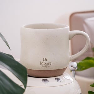 Può includere: Tazza in ceramica color crema con finitura maculata e base marrone. La tazza ha un manico curvo e il testo "Dr. Moore Est.2026" stampato. La tazza è appoggiata su un elettrodomestico color crema.