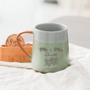 Puede incluir: Taza de cerámica verde claro y gris con la inscripción "Mr. & Mrs. Smith" y dos diseños de corazón. El asa es naranja. La fecha 00.10.2023 está impresa debajo. Un portavasos de madera está en el fondo.