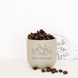 May include: A beige ceramic cup filled with dark roasted coffee beans. The cup features a mountain and moon design and the text "Bob's Espresso". Several coffee beans are scattered around the cup.