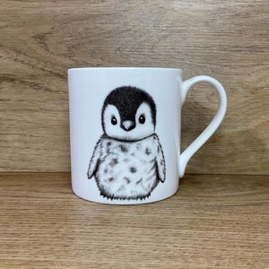 May include: White ceramic mug featuring a black and white illustration of a penguin chick. The penguin is facing forward with its wings tucked in and has a fluffy appearance.