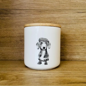 May include: White ceramic storage jar with a wooden lid. The jar has a black and white illustration of a dachshund wearing a tweed cap. The design is centered on the jar, which is cylindrical. The background is a light brown wooden surface.
