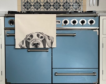 May include: A pale blue oven with stainless steel handles and knobs. A beige tea towel featuring a black and white dog portrait hangs on the oven door. Knives are mounted on the wall above the oven.