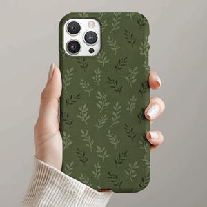 Puede incluir: Una funda de teléfono verde con un patrón repetido de pequeñas hojas estilizadas. La funda se sostiene en una mano con uñas cuidadas. La funda del teléfono tiene un acabado mate y un recorte para la cámara.