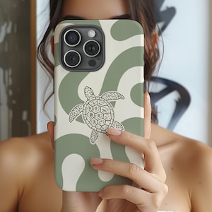 Puede incluir: Una funda para smartphone con un patrón abstracto ondulado en verde salvia y crema, que presenta una ilustración central en blanco y negro de una tortuga marina. La funda tiene un recorte para un sistema de cámara triple.