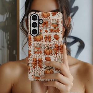 May include: A rectangular smartphone case featuring a repeating pattern of orange pumpkins, brown bows, maple leaves, and cups of coffee with whipped cream on a cream-coloured background with thin vertical stripes. The case has a black camera cutout.