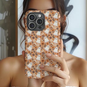 Puede incluir: Una funda para smartphone con un patrón repetitivo de pequeños fantasmas blancos con caras sonrientes, intercalados con flores naranjas, marrones y blancas y hojas de color verde oscuro sobre un fondo naranja claro.