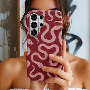Op de afbeelding: Een rechthoekig smartphonehoesje met een dieprode bordeauxrode achtergrond en een herhalend patroon van lichtroze, kronkelende slangenillustraties. Het hoesje heeft een uitsparing voor een camera met meerdere lenzen in de linkerbovenhoek.
