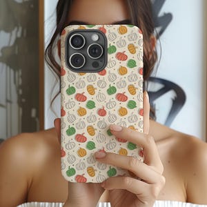 Puede incluir: Una funda para smartphone con un patrón repetitivo de pequeñas calabazas en tonos naranja, verde y blanco sobre un fondo crema. La funda tiene un gran recorte rectangular para la cámara en la parte superior izquierda, mostrando tres lentes.