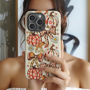 Puede incluir: Una funda de smartphone color crema con un patrón repetitivo de calabazas naranjas y blancas, lazos con estampado de leopardo marrón, pequeñas margaritas blancas y diminutas bellotas naranjas. El diseño cubre toda la parte trasera de la funda.