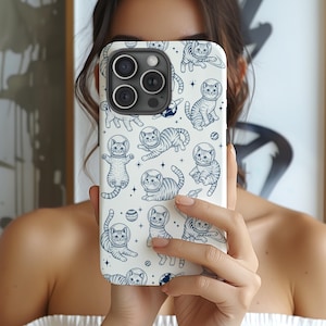 May include: White phone case featuring a repeating pattern of blue astronaut cats, planets, and stars. The cats are in various poses, wearing space helmets. The case has a camera cutout and is held by a person.