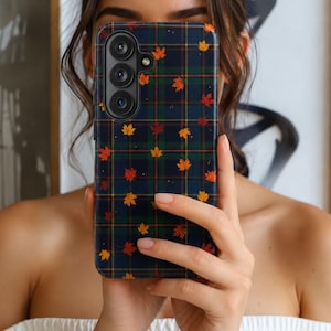 May include: A smartphone case featuring a dark navy blue, green, and yellow plaid tartan pattern decorated with small, scattered orange, red, and yellow maple leaves. The case has a vertical camera cutout on the upper left side with three lenses.