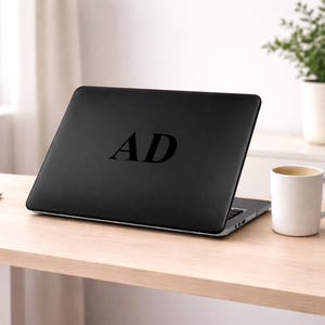 May include: A black laptop with the initials "AD" in a bold, sans-serif font on the lid. The laptop is open on a light-colored wooden desk, with a white mug and a small potted plant in the background.