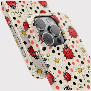 May include: A phone case with a cream background, featuring a pattern of red ladybugs, white daisies, and black and red polka dots. The case has a glossy finish and a camera cutout.