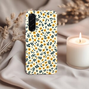 May include: A phone case with a floral pattern. The case features a cream-colored background adorned with small yellow flowers and dark blue leaves. The phone case has a camera module on the upper left side.