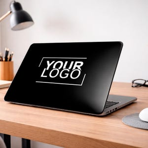 Op de afbeelding: Zwarte laptop met de witte tekst "YOUR LOGO" in een witte rechthoekige lijst. De laptop staat open op een houten bureau, met een bureaulamp en kantoorbenodigdheden op de achtergrond.