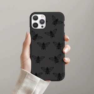Puede incluir: Funda de teléfono negra con un patrón repetido de ilustraciones detalladas de abejas. La funda se sostiene en una mano, mostrando el diseño. Las abejas son oscuras, contrastando con el negro de la funda.