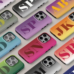 Puede incluir: Una colección de fundas para teléfonos inteligentes en varios colores, cada una con un monograma de dos letras. Las fundas tienen un acabado brillante y están dispuestas sobre una superficie gris claro. Los colores incluyen morado, rosa, verde y azul.