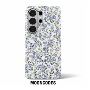 May include: A white phone case with a floral pattern of small blue flowers and green leaves. The case has a camera module with three lenses and a flash. The brand name "MOONCODES" is printed in black at the bottom.