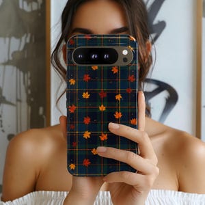 May include: A smartphone case featuring a dark blue and green plaid tartan pattern overlaid with small, scattered orange and red maple leaves. The case has a horizontal camera cutout at the top, revealing three circular lenses and a flash component.