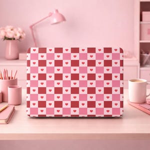 Puede incluir: Una funda para portátil con un patrón de tablero de ajedrez rosa y rojo, cada cuadrado contiene un pequeño corazón rojo. La funda está sobre un escritorio de color claro, con una lámpara rosa y otros accesorios rosas en el fondo.
