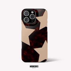 Puede incluir: Una funda para smartphone con un patrón de carey en marrón oscuro y negro, superpuesto con grandes estrellas beige sólidas. La funda tiene un recorte para un sistema de cámara de tres lentes en la parte posterior. El texto MOONCODES HOUSTON, TX aparece debajo.
