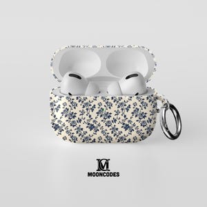 Puede incluir: Una funda abierta para AirPods, de color crema, con un estampado floral azul. Contiene unos AirPods blancos. Un mosquetón plateado está sujeto al lateral. El texto "MOONCODES" está impreso en negro en la parte inferior.