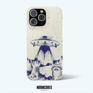 Puede incluir: Una funda de teléfono color crema con una ilustración gráfica azul oscuro de un platillo volante proyectando luz sobre una escena forestal con dos mapaches. Un mapache está de pie con los brazos levantados hacia el OVNI, el otro mira al frente.