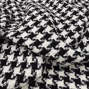 May include: Close-up of a black and white houndstooth fabric. The woven material has a repeating pattern of black shapes on a white background. The fabric is folded, creating shadows and texture.