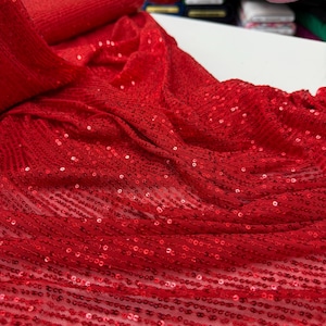 Peut inclure: Un tissu rouge vif orné d'un motif dense de petites paillettes circulaires disposées en rangées linéaires. Le matériau possède une base en maille semi-transparente qui permet à la couleur rouge éclatante de capter la lumière, créant un aspect texturé.