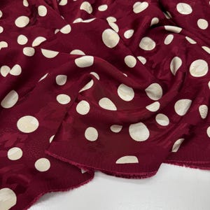 a close-up view of a fabric with a maroon color and white polka dots. The fabric appears to be made of silk or a similar material, and the polka dots are scattered across the fabric in a pattern. The fabric has a slightly wrinkled texture, giving it a casual and relaxed appearance.