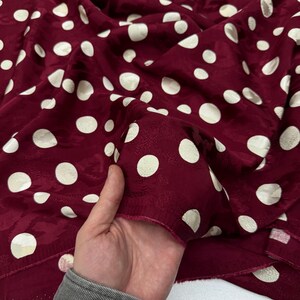 A hand is holding a fabric with a maroon color and white polka dots, which appears to be a close-up view of the fabric.