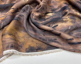 May include: Close-up of a fabric with a brown, black, and gold abstract pattern. The fabric is draped, showing folds and textures. The edge of the fabric has a white fringe.