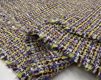 Op de afbeelding: Close-up van een kleurrijke tweed stof met een geweven patroon. De stof heeft een mix van kleuren, waaronder geel, paars, bruin en blauw. De randen van de stof zijn gerafeld, wat textuur en detail aan het materiaal toevoegt.
