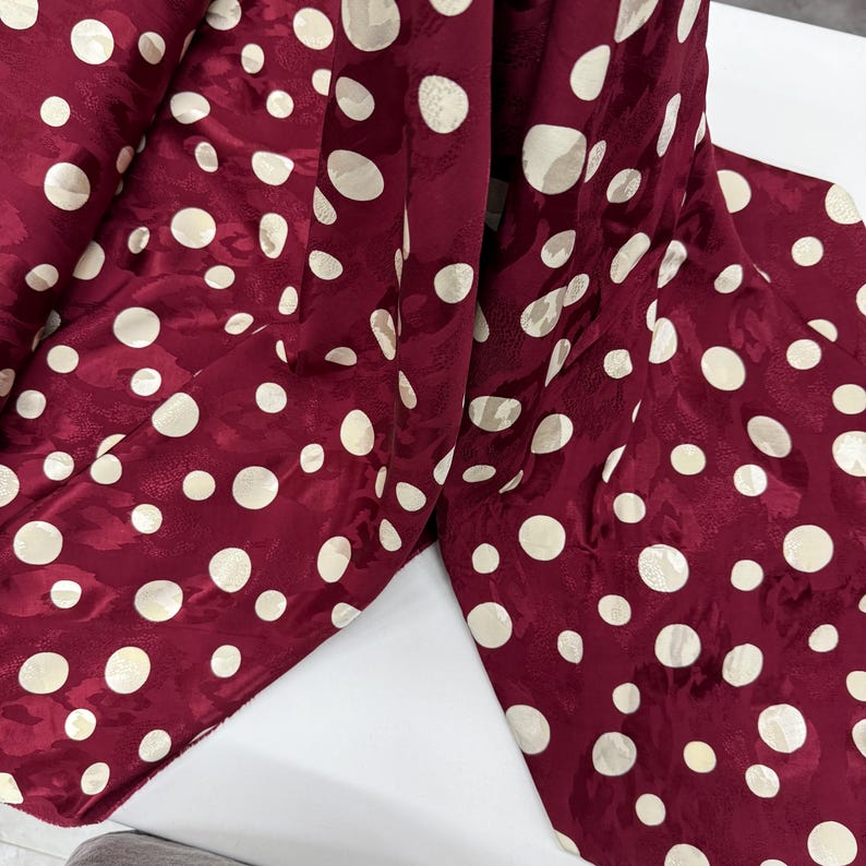 a close-up view of a fabric with a maroon color and white polka dots. The fabric appears to be a piece of clothing or textile, with the polka dots covering the entire surface.