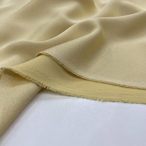 Puede incluir: Primer plano de una tela de color amarillo pálido con una superficie ligeramente texturizada. La tela está doblada, mostrando el revés de la tela, que es de un tono amarillo más claro.