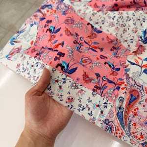 May include: A patchwork fabric quilt featuring a repeating pattern of pink, white, and blue floral and paisley motifs. The textile is composed of rectangular segments stitched together, displaying a vibrant mix of botanical illustrations and intricate swirls.