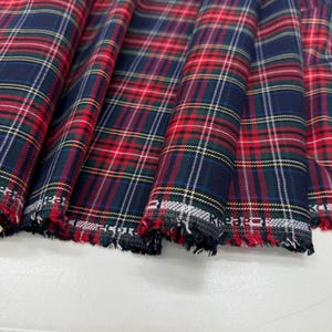 Può includere: Primo piano di un tessuto a quadri piegato. Il tessuto presenta un classico motivo tartan con linee rosse, blu navy, verdi e bianche che si intersecano. I bordi del tessuto sono sfilacciati, rivelando la trama grezza del materiale.
