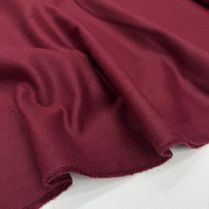 May include: Close-up of a deep burgundy fabric with a soft, textured surface. The fabric is draped, showing folds and a raw edge. The color is a rich, saturated shade of red with a hint of brown.