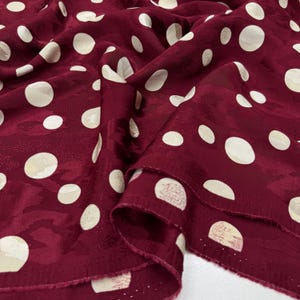 a close-up view of a fabric with a maroon color and white polka dots. The fabric appears to be made of silk or a similar material, and the polka dots are scattered across the fabric in a pattern.