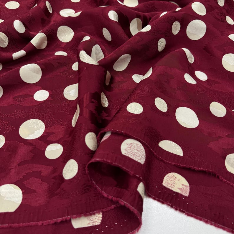 a close-up view of a fabric with a maroon color and white polka dots. The fabric appears to be made of silk or a similar material, and the polka dots are scattered across the fabric in a pattern.