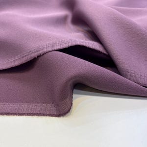 Könnte beinhalten: Nahaufnahme eines weichen, mauvefarbenen Stoffes mit einer subtilen Textur. Der Stoff ist umgeklappt und zeigt die rohe Kante und eine genähte Naht.