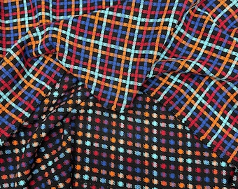 Puede incluir: Tela negra con un colorido estampado de cuadros en rojo, naranja, azul y azul claro. La tela está doblada, revelando un patrón de puntos en la parte inferior en colores similares. La tela parece ser un material tejido.