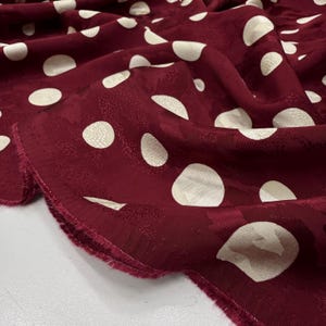 a close-up view of a fabric with a maroon color and white polka dots. The fabric appears to be made of a soft, plush material, possibly a type of chiffon or a similar textile. The polka dots are scattered across the fabric, creating a playful and festive pattern. The fabric is folded and crumpled, giving it a casual and relaxed appearance.
