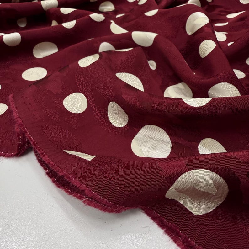 a close-up view of a fabric with a maroon color and white polka dots. The fabric appears to be made of a soft, plush material, possibly a type of chiffon or a similar textile. The polka dots are scattered across the fabric, creating a playful and festive pattern. The fabric is folded and crumpled, giving it a casual and relaxed appearance.